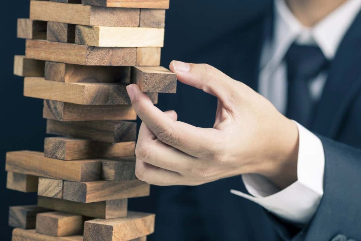 man in business suit pulls a piece out of wobbly wooden tower
